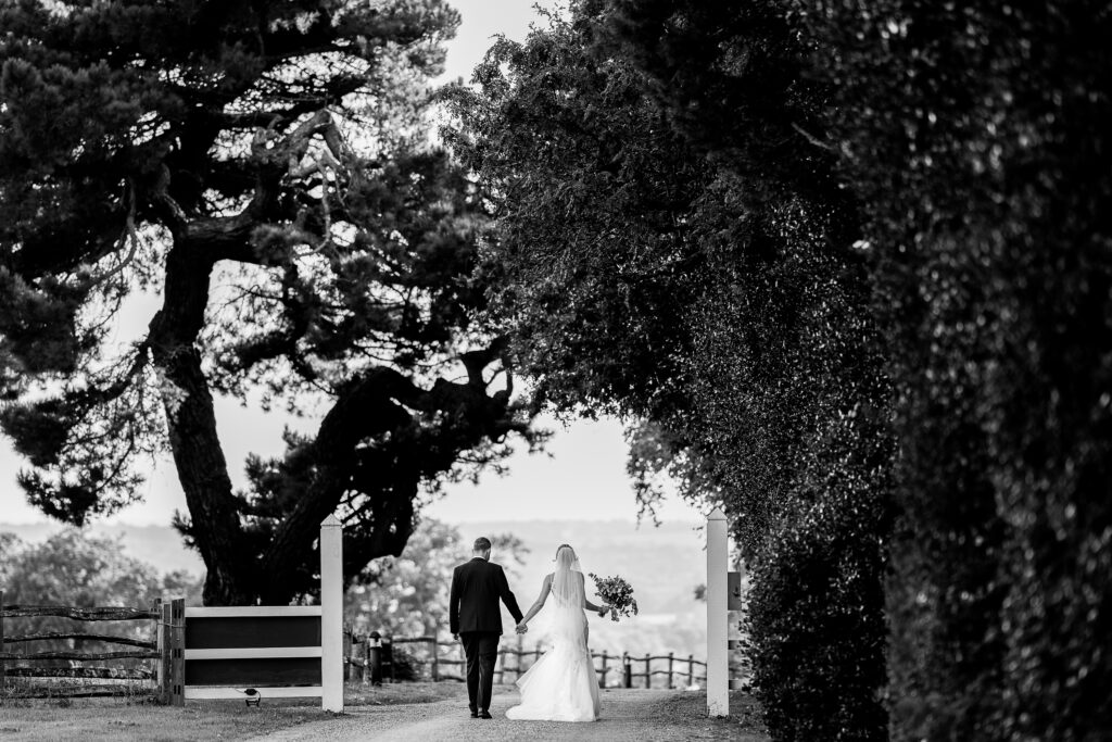 A black-and-white image of a bride and groom holding hands while walking down a tree-lined path, with the bride wearing a long dress and veil, and the groom in a suit. They are framed by trees and a fence in the background.