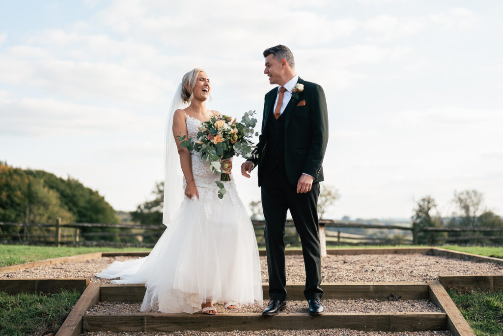 A bride and groom stand outdoors on wooden steps. The bride, in a white gown, holds a bouquet and is laughing. The groom, in a dark suit with an orange tie, smiles at her. They are surrounded by a natural landscape with trees and a cloudy sky.