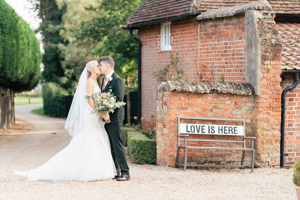 A bride and groom share a kiss in front of a rustic brick building with a sign on a bench that reads "LOVE IS HERE." The bride is in a white gown, and the groom is in a black suit. Lush greenery surrounds the scene.