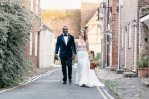 A bride and groom walk hand in hand down a cobblestone street lined with brick buildings. The groom wears a dark suit and bow tie, while the bride is in a white gown, holding a colorful bouquet.