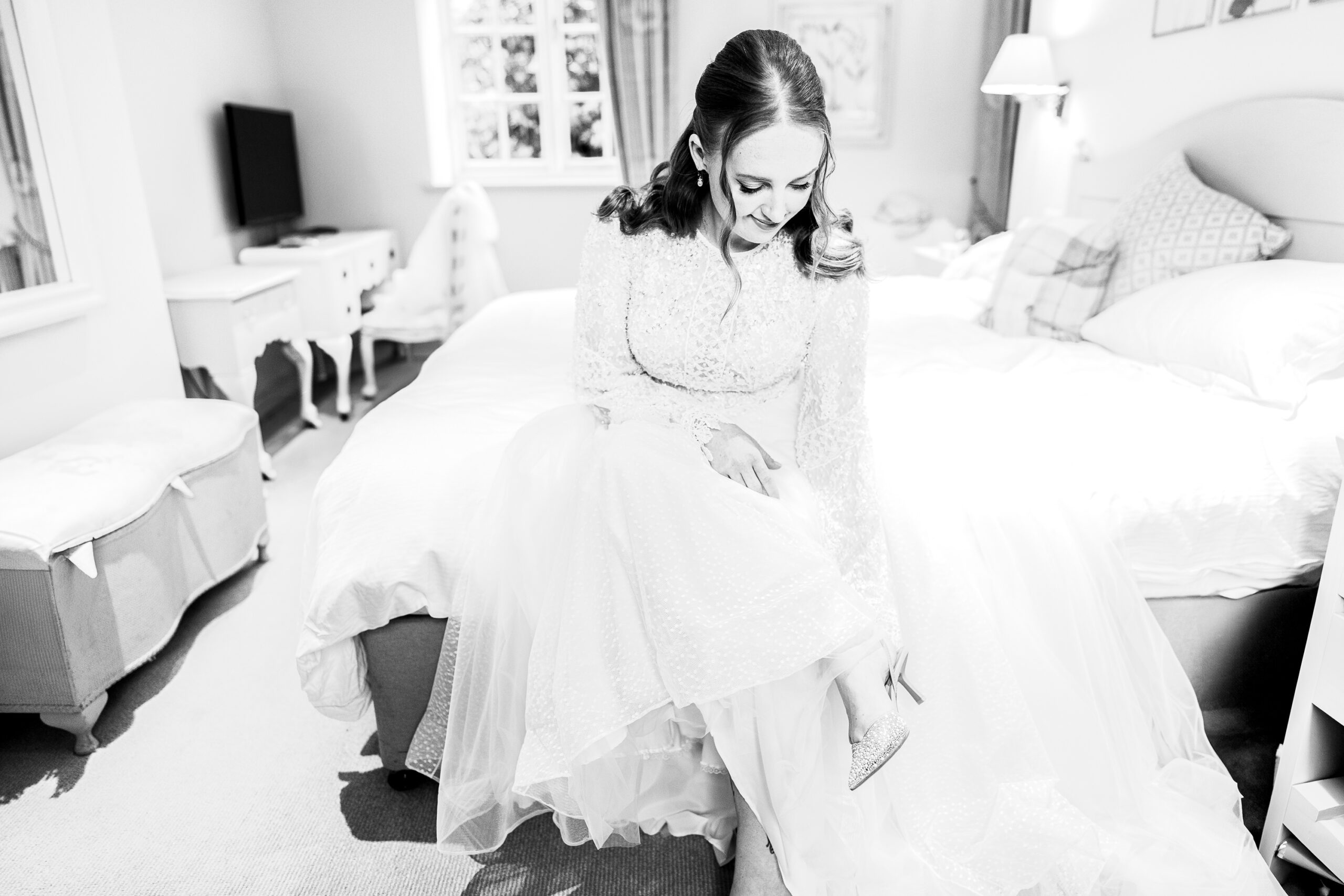 A bride in a white wedding dress sits on the edge of a bed in a bright room, adjusting her shoe. The room features a bed, a window, and various decorative items, creating a serene and elegant atmosphere.