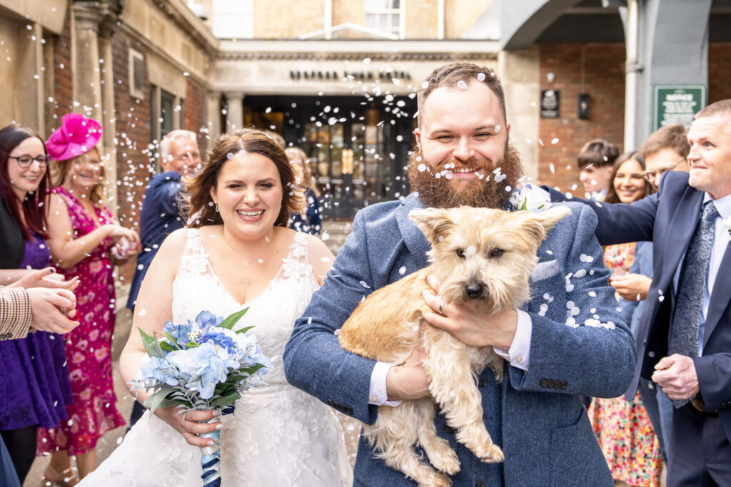 A joyful couple exits a building after their wedding ceremony. The bride holds a bouquet of blue flowers, while the groom carries a small dog. Guests cheer and toss confetti. The scene is filled with celebration and smiles. Bedford Swan Hotel & Spa, Bedford.