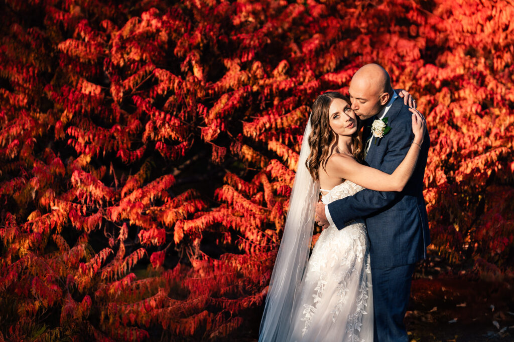 A bride and groom embrace in front of vibrant red and orange foliage. The bride wears a white lace gown and veil, while the groom is in a dark suit with a boutonnière. The lighting highlights the couple and the colorful autumn background.