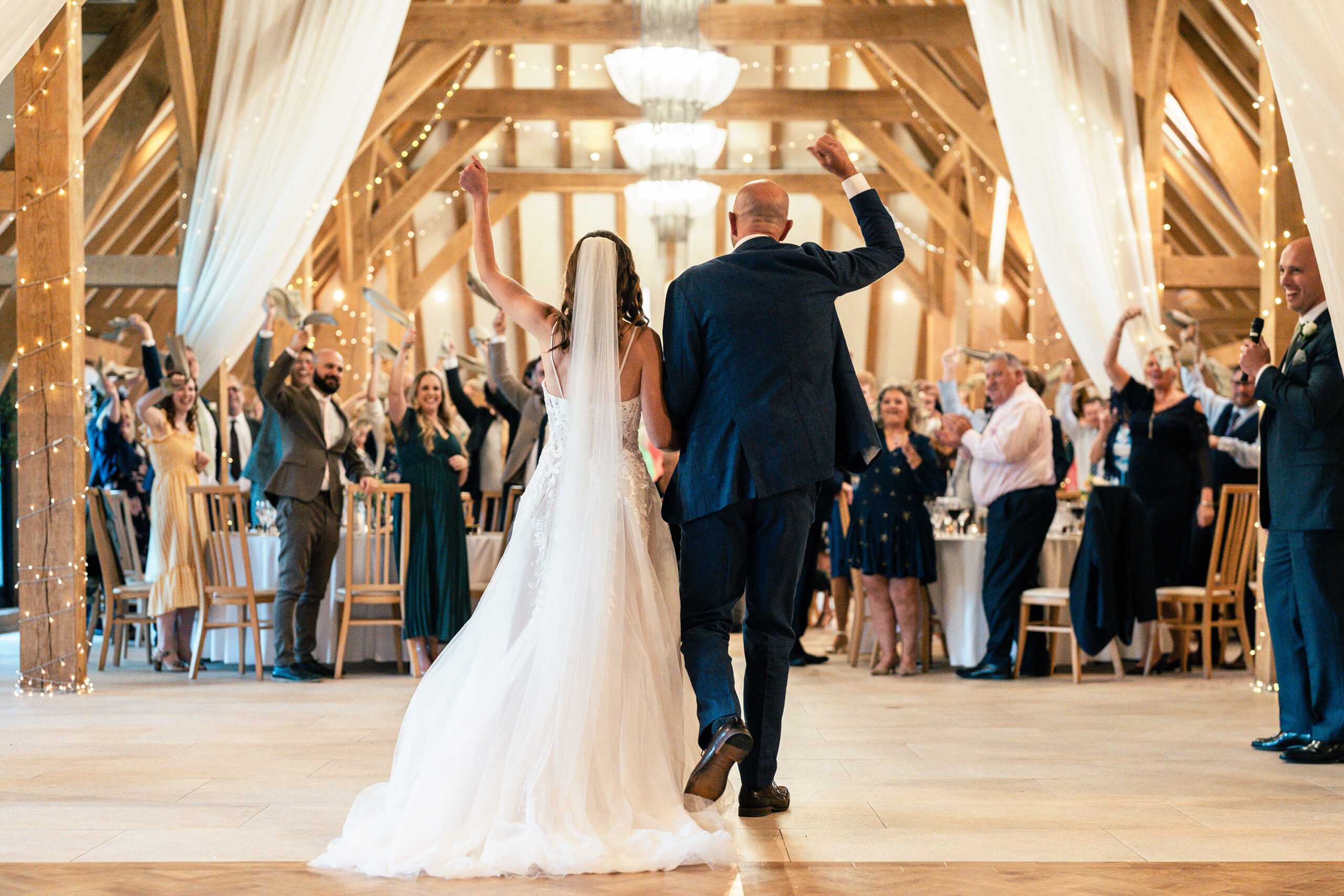 A newlywed couple, the bride in a white gown and the groom in a blue suit, enters a warmly lit reception hall. Guests seated at tables applaud and cheer, surrounded by rustic wooden beams and draped white fabric with string lights. Old Kent Barn, Kent