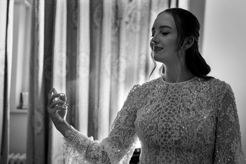 A bride in a detailed lace dress sprays perfume, with a slight smile. She stands in front of curtains, creating an elegant and serene atmosphere. The image is in black and white, highlighting the texture of the fabric and the soft mist of the spray.