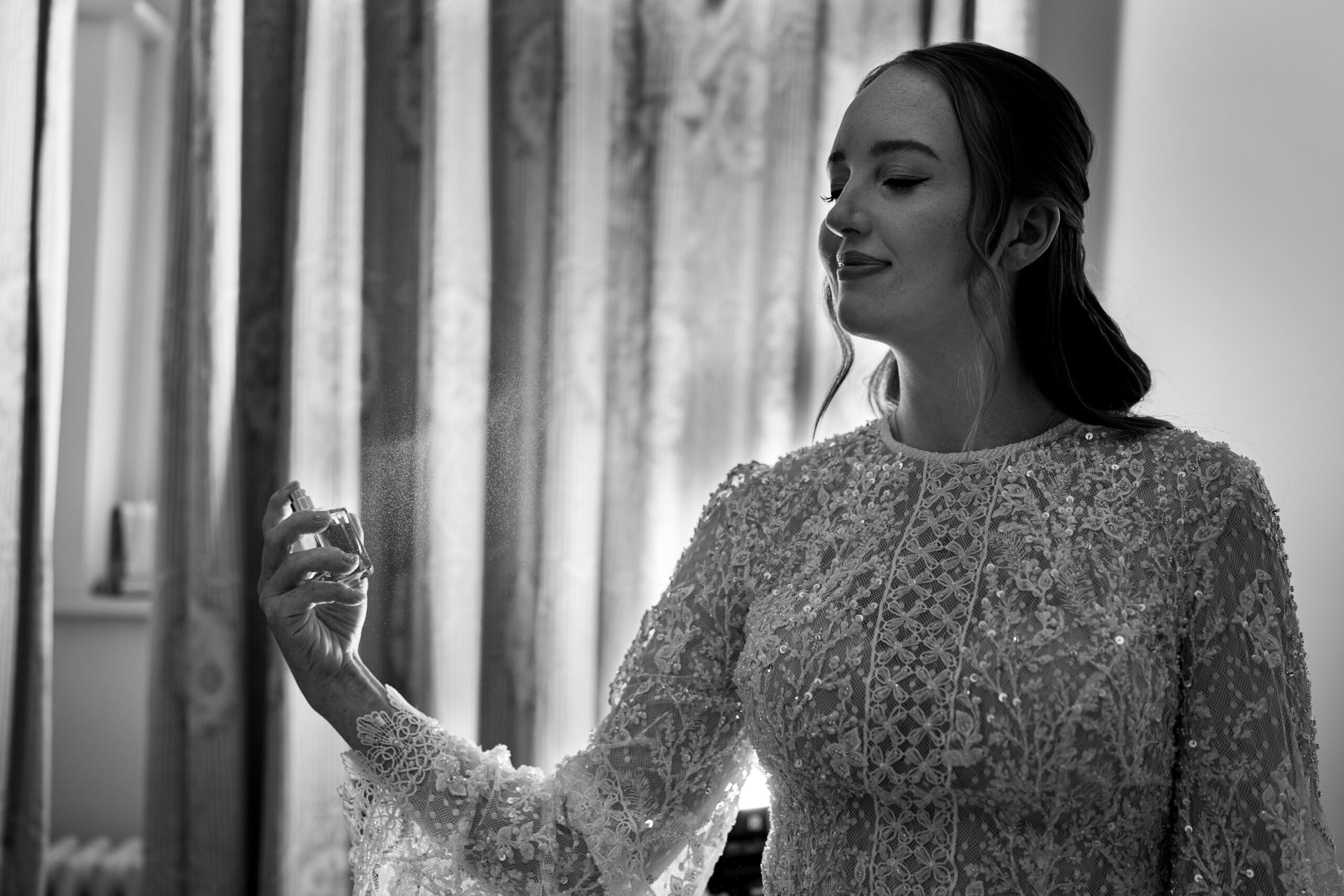 A bride in a detailed lace dress sprays perfume, with a slight smile. She stands in front of curtains, creating an elegant and serene atmosphere. The image is in black and white, highlighting the texture of the fabric and the soft mist of the spray.