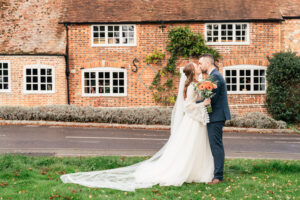 A bride in a white dress and a groom in a dark suit kiss in front of a brick house with white-framed windows. The bride holds a bouquet of orange and white flowers. They stand on a grassy area beside the road.