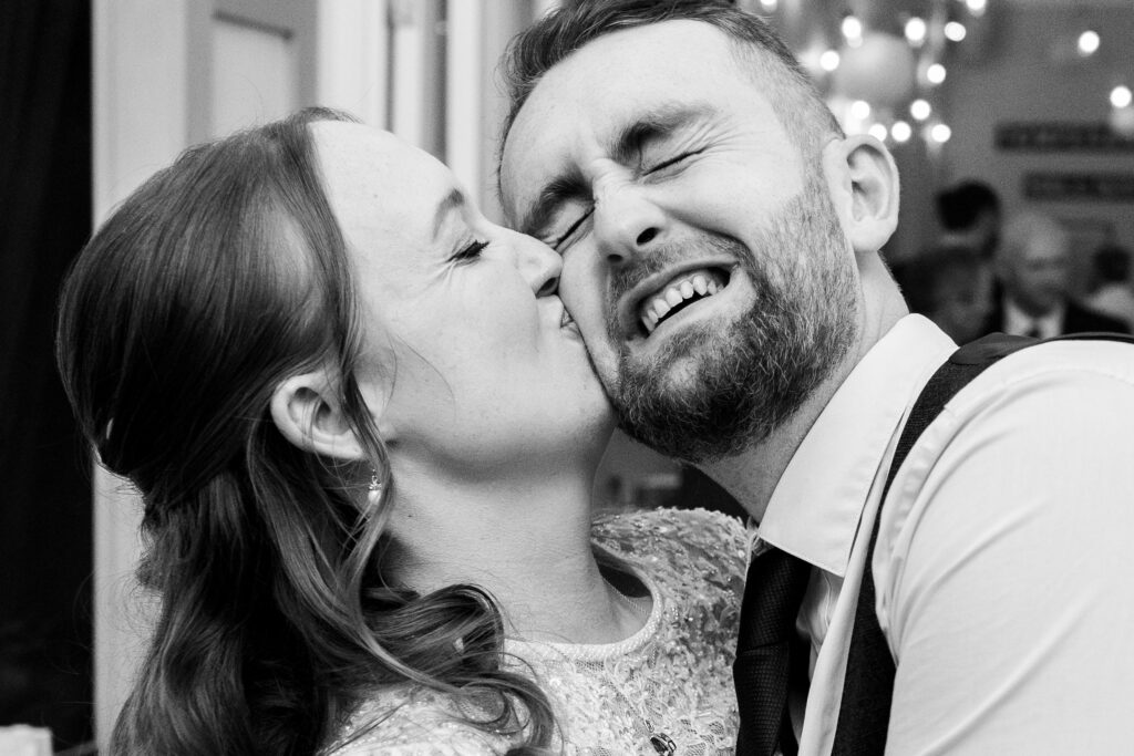 A black and white photo of a woman kissing a smiling man on the cheek. Both appear joyful, and the background shows softly lit hanging lights, creating a festive atmosphere.