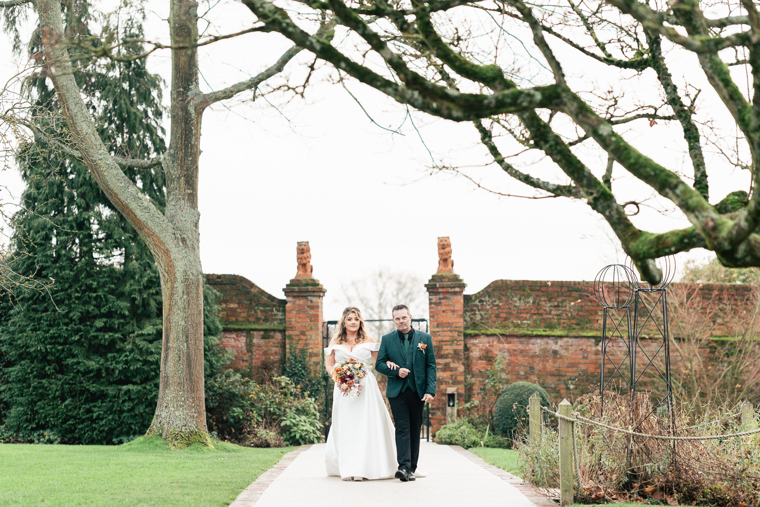 A bride walks down the long walk at Gaynes Park, in a white gown holding a bouquet walks with her father. They are under leafless trees, with a brick wall and lush greenery in the background.