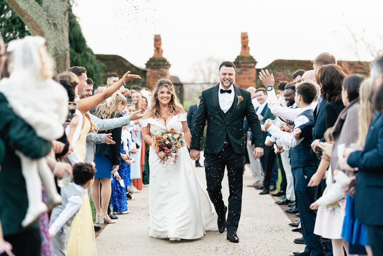 A bride and groom in wedding attire walk dow the long walk at Gaynes Park wedding venue, hand in hand down an outdoor aisle as guests cheer and throw confetti. The couple looks joyful, surrounded by friends and family in a festive atmosphere, with trees and structures in the background.