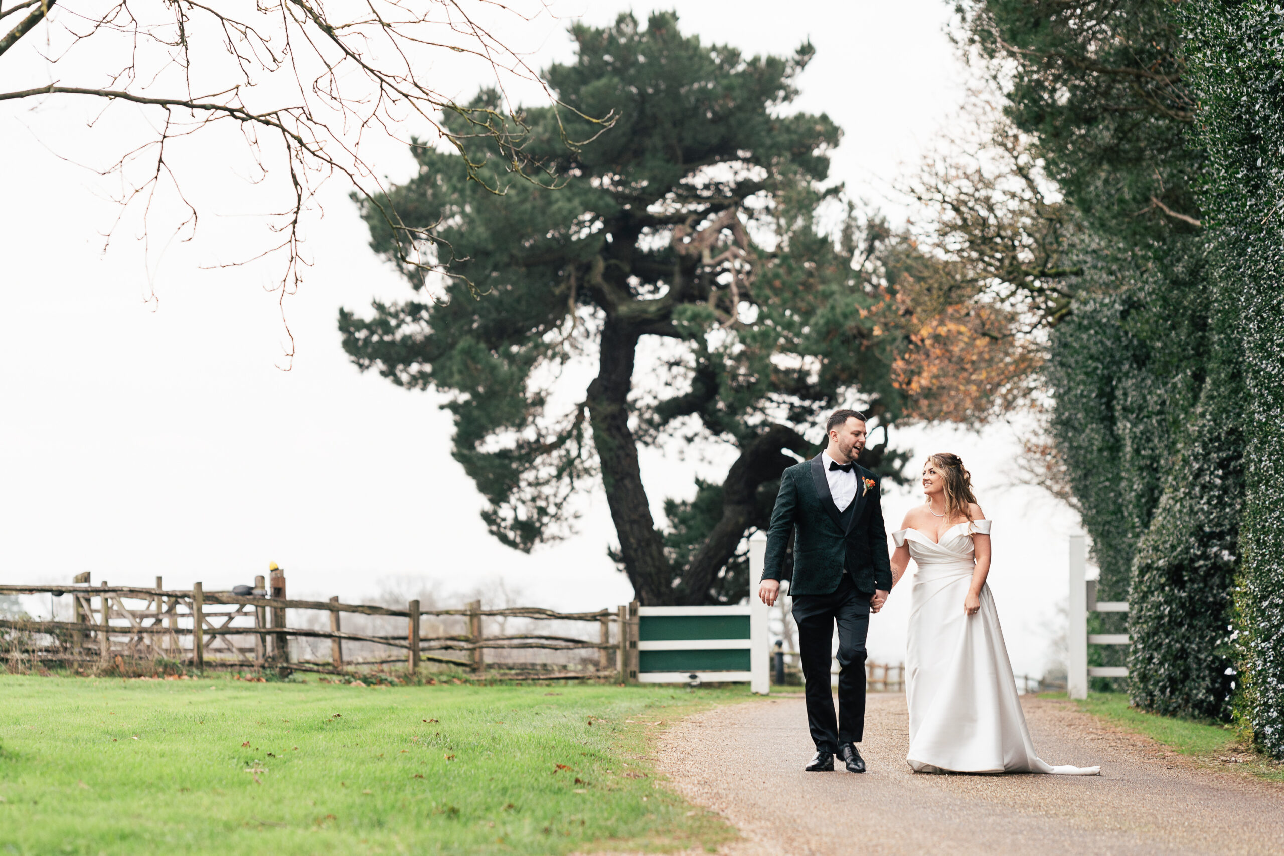 A bride and groom walk hand in hand along the Gaynes Park driveway, lined with greenery and a large tree in the background. The bride wears a white gown, and the groom is in a dark suit and bow tie. The scene is serene and picturesque.