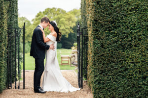 A bride and groom embrace in a garden pathway at Hedsor House. Buckinghamshire, framed by tall, manicured hedges. The woman wears an ivory off-the-shoulder wedding gown, and the man wears a dinner suit. They stand closely, sharing an intimate moment under an open wrought iron gate.