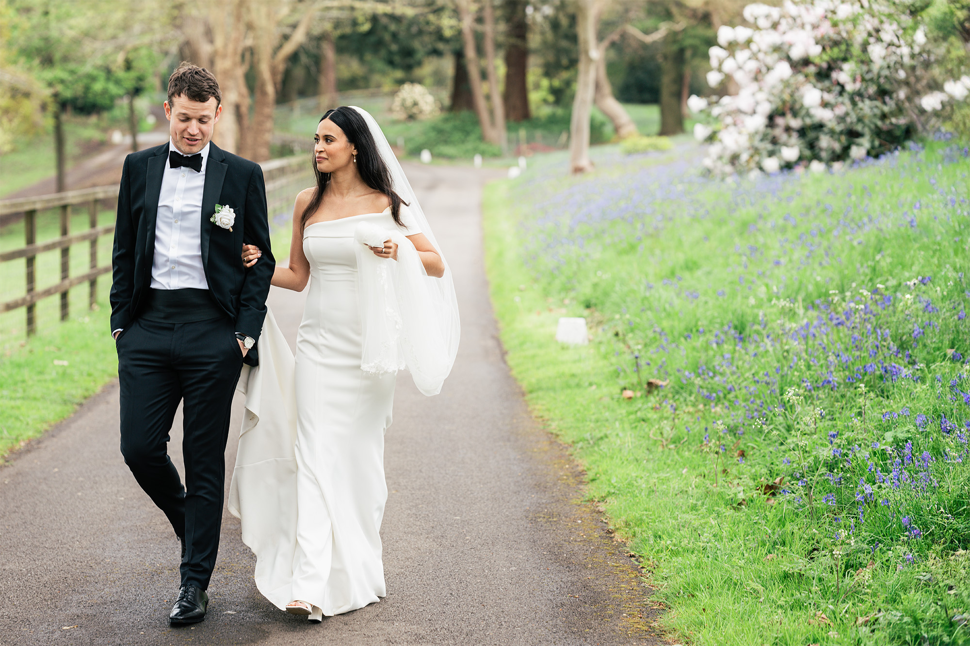 A bride in a white dress and a groom in a black suit walk hand in hand down a paved path, surrounded by lush greenery and purple flowers. They appear to be enjoying a peaceful moment together on their wedding day.