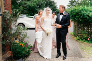 A bride in a white gown and veil holds a bouquet of white flowers while walking with her father in a black tuxedo. Three bridesmaids in matching dresses stand nearby. A white vintage rolls Royce car is parked in the background, surrounded by greenery and flowers. They are about to enter a Greek Orthodox Church.