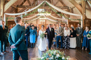 A bride in a white gown walks down the aisle at Lains Barn, with her father in this rustic barn venue, adorned with greenery and draped fabric. Guests stand in anticipation. The groom is visible from the back, wearing a dark green suit.