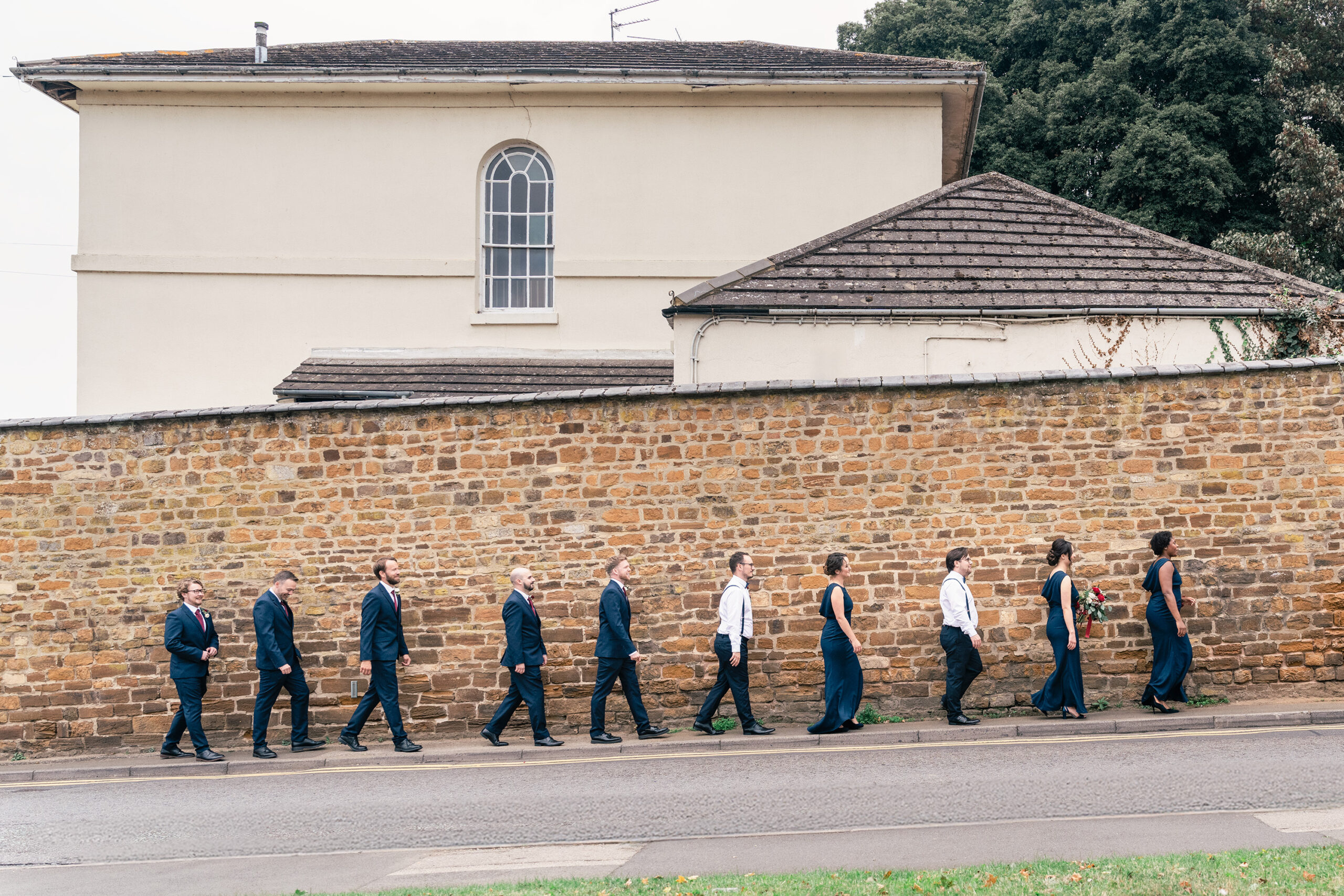 A group of people in formal wedding attire, walk single file along a brick wall, with a beige building and trees in the background.