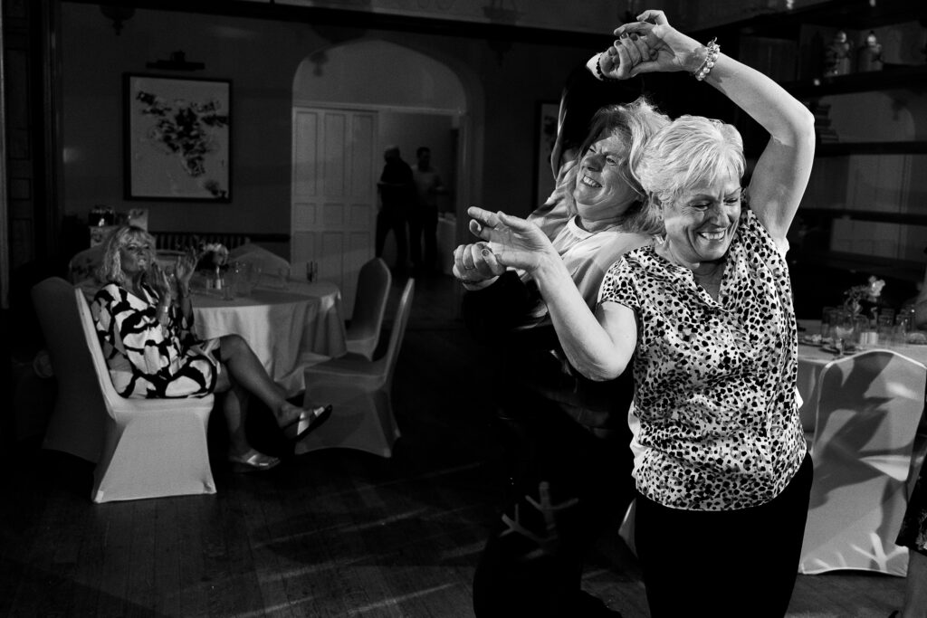 Two women joyfully dancing together in a warmly lit room, smiling broadly. A woman in a patterned dress sits at a table in the background, watching them. The atmosphere is lively and celebratory.