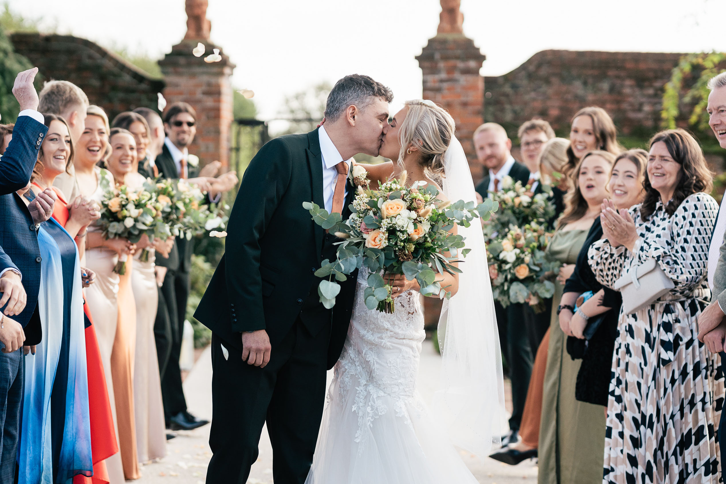 A bride and groom share a kiss surrounded by cheering guests at an outdoor wedding. The bride holds a bouquet of flowers and wears a white dress with a veil, while the groom is in a dark suit. Guests are clapping and smiling in celebration.