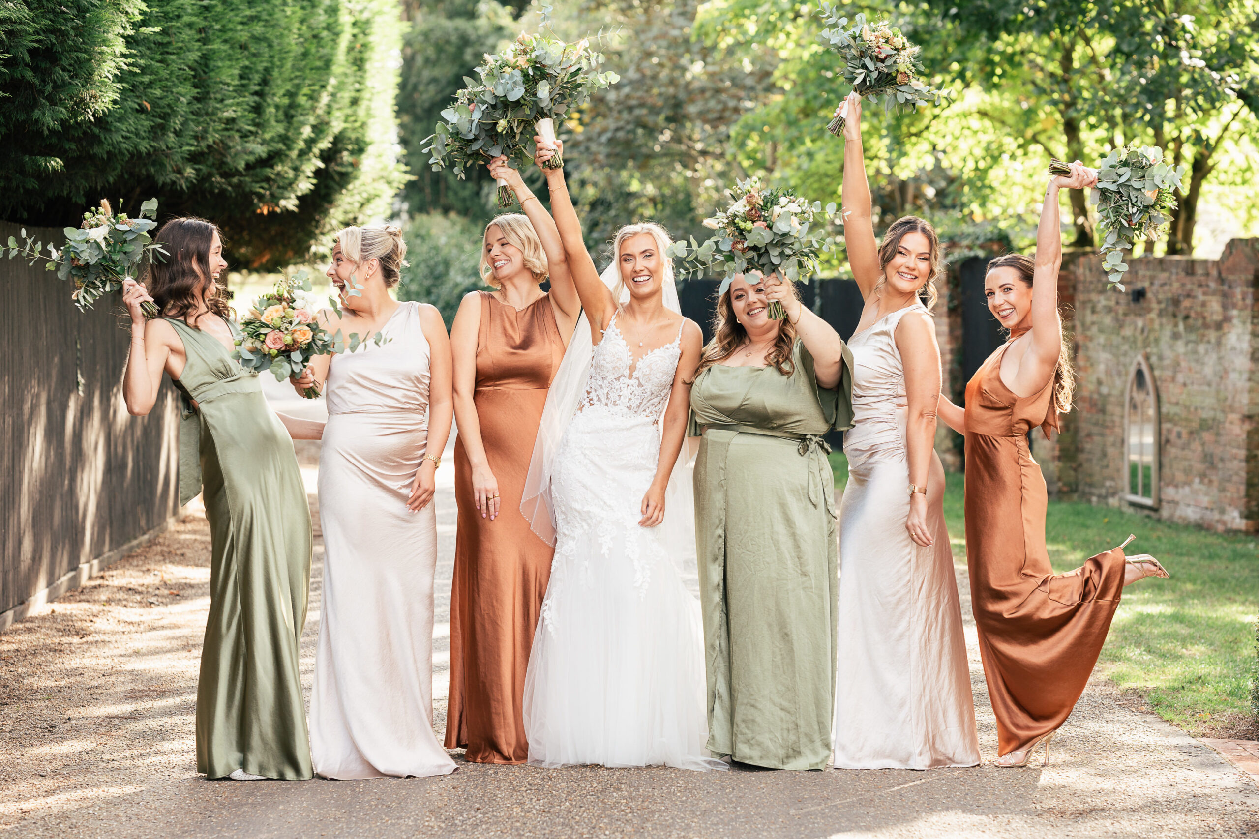 A group of seven women stand in a garden, wearing elegant dresses in shades of green, beige, and copper. They are smiling and holding bouquets, with trees and a brick wall in the background.