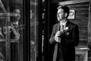 A man in a suit adjusts his tie while looking out of a window. His reflection is visible in the glass. He has a boutonniere on his lapel, and the setting has wooden walls, suggesting a rustic location. The image is in black and white.