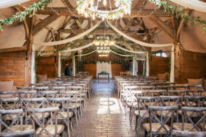 A rustic wedding venue with wooden beams and an aisle lined with wooden chairs. White fabric and greenery drape from the ceiling. A chandelier hangs above, and light streams through a window onto the brick floor.