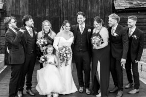 A black and white photo of a wedding party. The bride and groom stand in the center, surrounded by groomsmen, bridesmaids, and a flower girl. All are smiling and dressed in formal attire, standing in front of a rustic wooden backdrop.