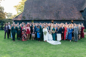 A large group of people, dressed in formal attire, pose for a group photo outside. The bride and groom stand in the center, with the bride holding a bouquet. They are in front of a rustic wooden building on a sunny day.