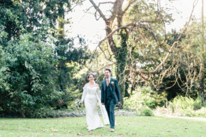 A bride and groom walk hand in hand on a grassy area surrounded by trees. The bride wears a white dress and the groom is in a dark suit with a boutonniere. Sunlight filters through the foliage, creating a serene and joyful setting.