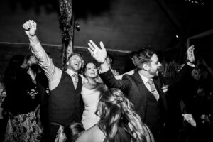 A black-and-white photo of a joyful wedding celebration. The bride, wearing a gown, stands between two men in suits, all with arms raised in enthusiasm. Guests surround them, some partially visible, adding to the lively atmosphere.