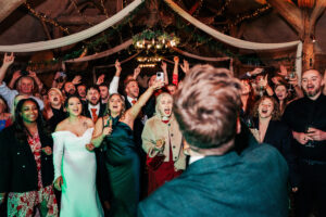 A lively group of people at a party, some with drinks in hand, all facing forward with expressions of joy and celebration. They are captured mid-cheer under a rustic ceiling adorned with draped fabric and string lights.