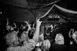 A black and white photo of a lively crowd at an indoor event. People are standing and cheering, with many raising their hands. Decorations and lights hang from the ceiling, and a person on a microphone engages the audience.