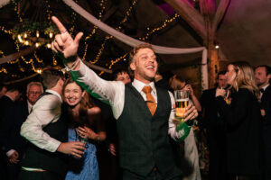 A lively wedding reception scene with a joyful group of people dancing. A man in a vest and tie holds a drink and gestures energetically. Others are smiling and celebrating under warm, festive lighting and decorative drapes.