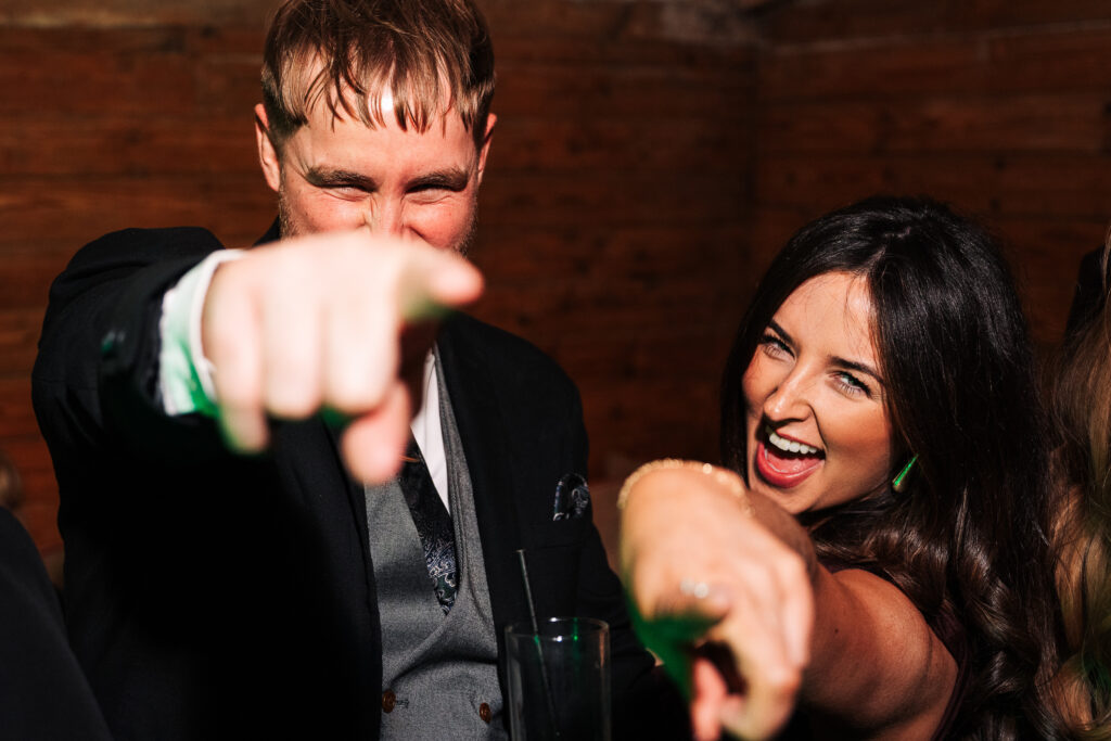 A man in a suit and a woman in a black dress are smiling and pointing playfully toward the camera. They appear to be enjoying themselves at an indoor event with wooden walls in the background.