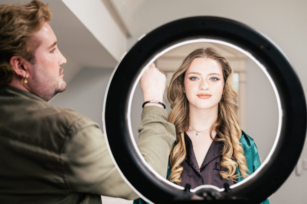 A makeup artist adjusts the hair of a woman with long, wavy locks, as she sits behind a circular ring light. The woman looks directly at the camera, wearing a teal robe, and the setting appears to be indoors.