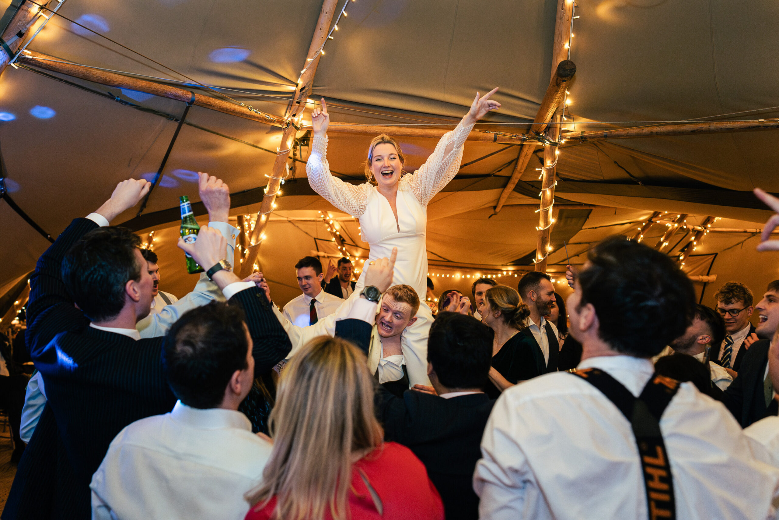 A joyful scene at a party inside a tent with string lights. A woman in a white dress is lifted into the air by a group of cheering people. Everyone is dressed in formal attire, celebrating with smiles and raised hands.