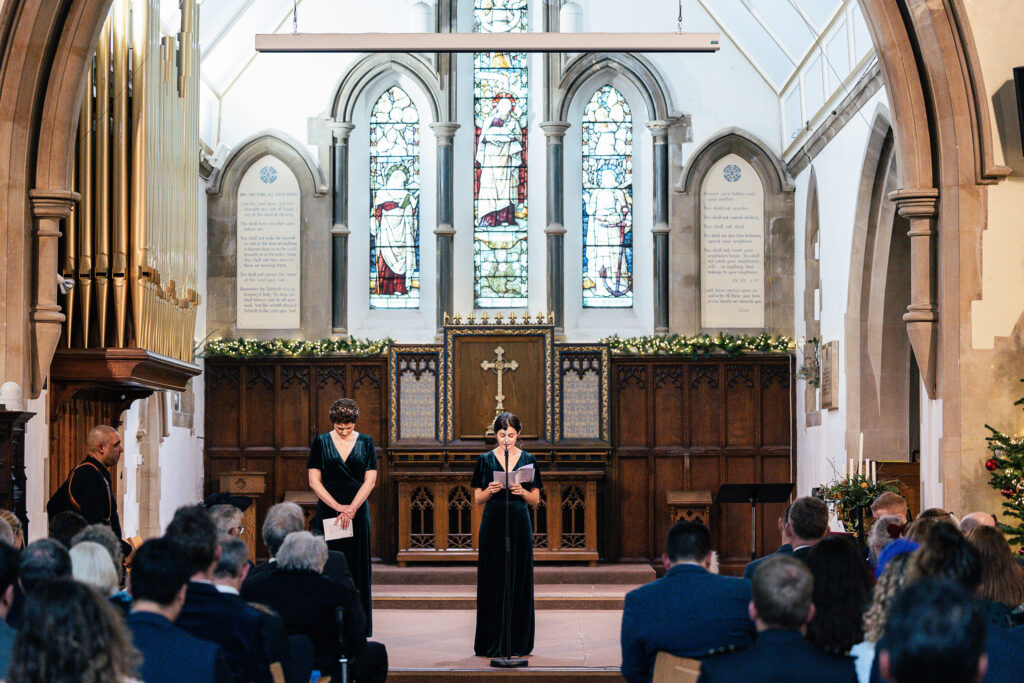 Two people stand at the front of a church with stained glass windows and wooden pews. One is speaking, and the other is holding a piece of paper. The audience is seated, facing them. The church interior is lit with natural light.