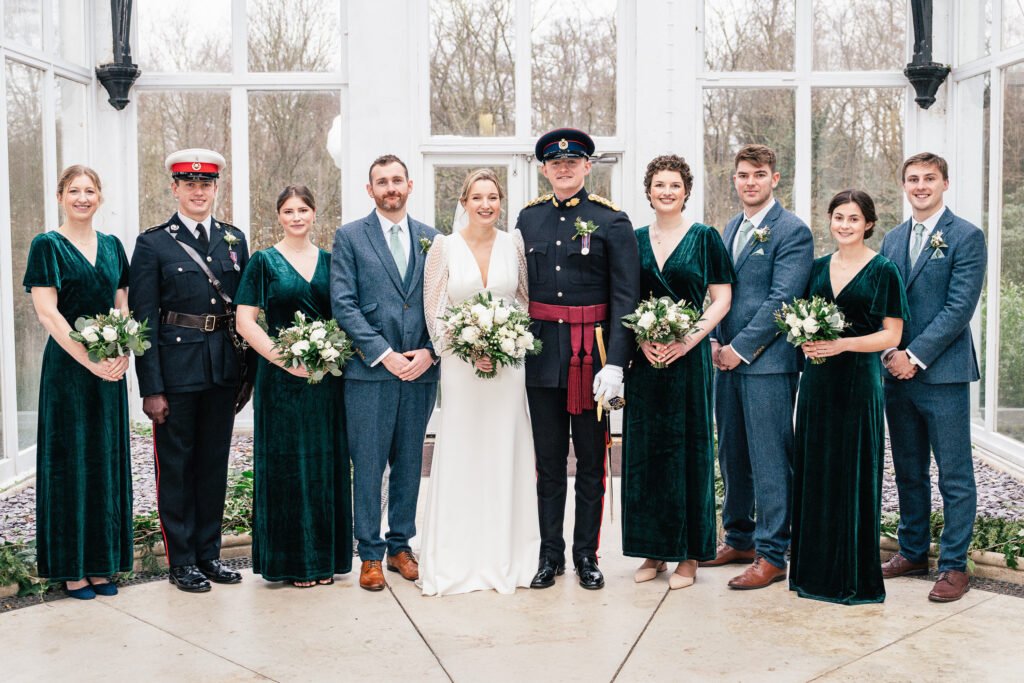 A wedding party poses indoors. The bride and groom are at the center, flanked by bridesmaids in green dresses and groomsmen in gray suits. Two men in military uniforms are at either end of the group, all holding bouquets or wearing boutonnieres.