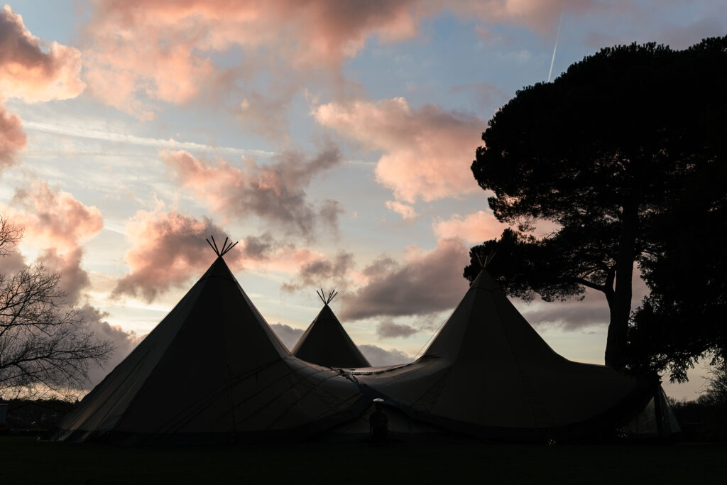 Three large tent structures silhouetted against a dramatic sky with pink and gray clouds at sunset. A tree stands to the right, and branches are visible on the left, creating a serene and atmospheric scene.