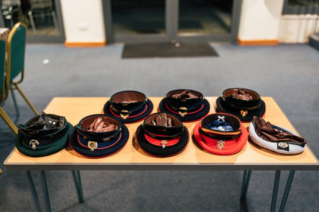 A table displays several military-style hats, each with a different colored brim and detailed emblem. The hats are arranged neatly in two rows of four on a light wood table in a carpeted room. Chairs and part of a door are visible in the background.