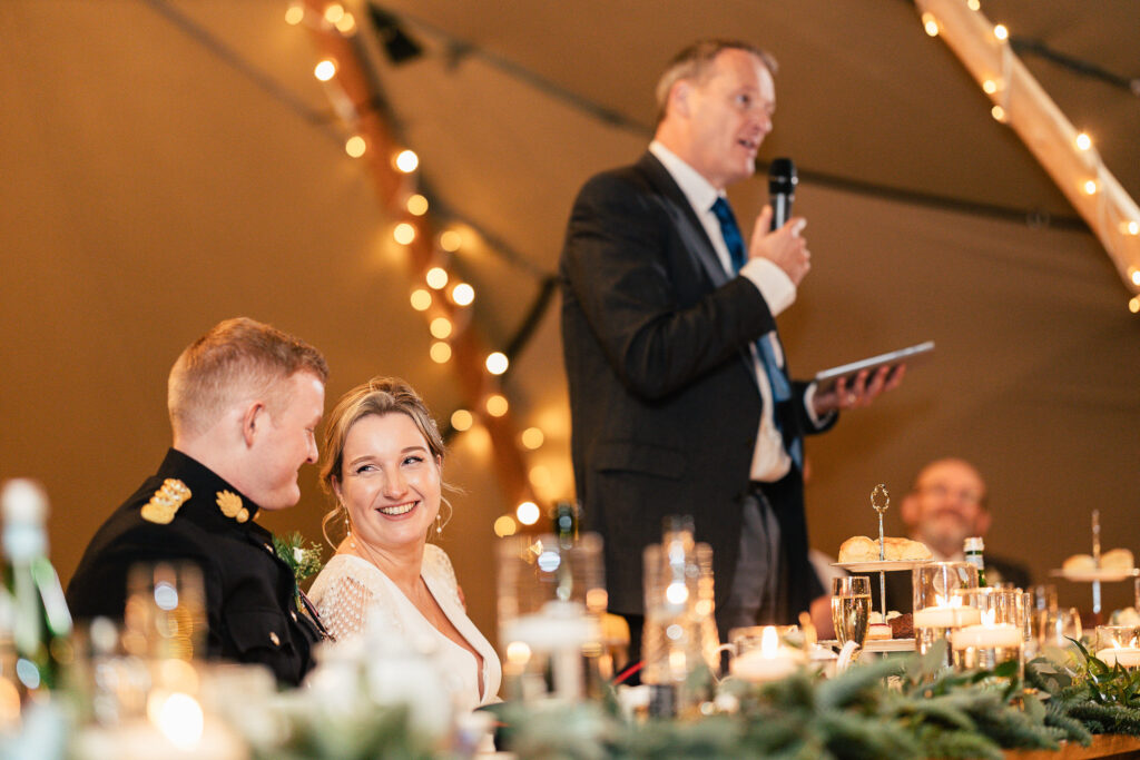 A woman in a wedding dress smiles at a man in a military uniform at a decorated reception table. A man in a suit stands to speak into a microphone, holding a tablet. Warm string lights hang above, creating a festive atmosphere.