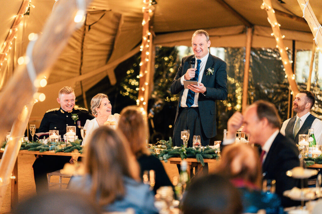 A man in a suit stands smiling, holding a microphone, giving a speech at a wedding reception. The bride and groom are seated nearby, also smiling. The tent is decorated with string lights, candles, and greenery. Guests sit around tables listening.