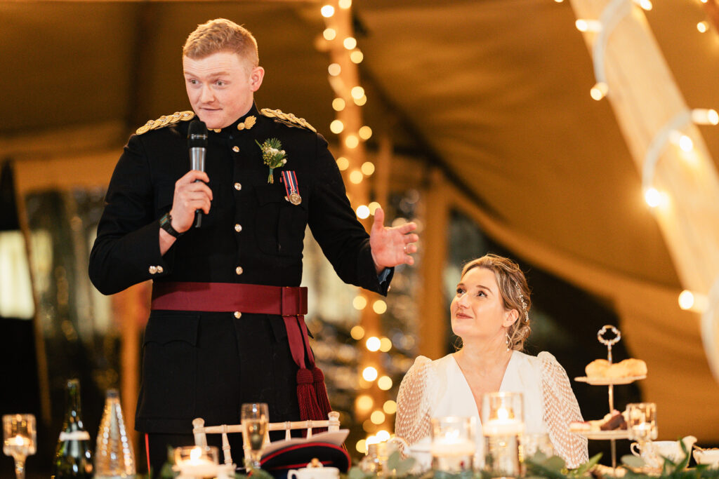 A person in a military dress uniform holds a microphone and speaks at a decorated table adorned with flowers and candles. A seated woman in a white dress looks up at them, with string lights creating a warm ambiance in the background.