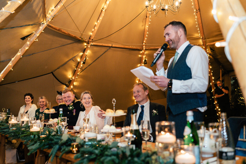 A man in a vest and tie speaks into a microphone, smiling, at a warmly lit event inside a tent. Guests, seated at a table adorned with candles and greenery, listen and smile. The ambiance is casual and festive.