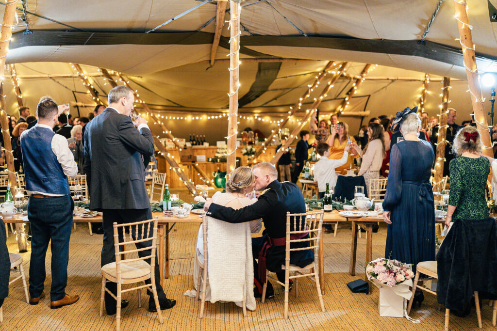 A couple sits closely at a wedding reception in a tent decorated with string lights. Guests toast around them, some standing and others seated at wooden tables. The atmosphere is warm and festive.