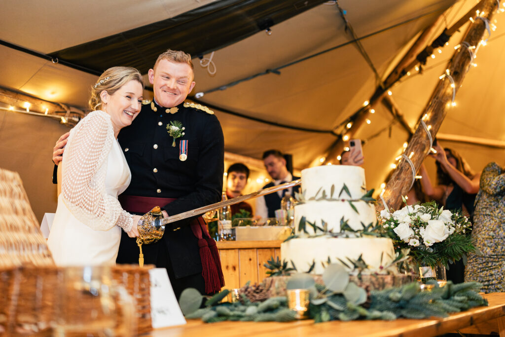 A bride and groom smile while cutting a tiered wedding cake with a sword. The bride wears a white dress, and the groom is in a dark formal military uniform. They are in a warmly lit tent decorated with string lights, surrounded by guests.