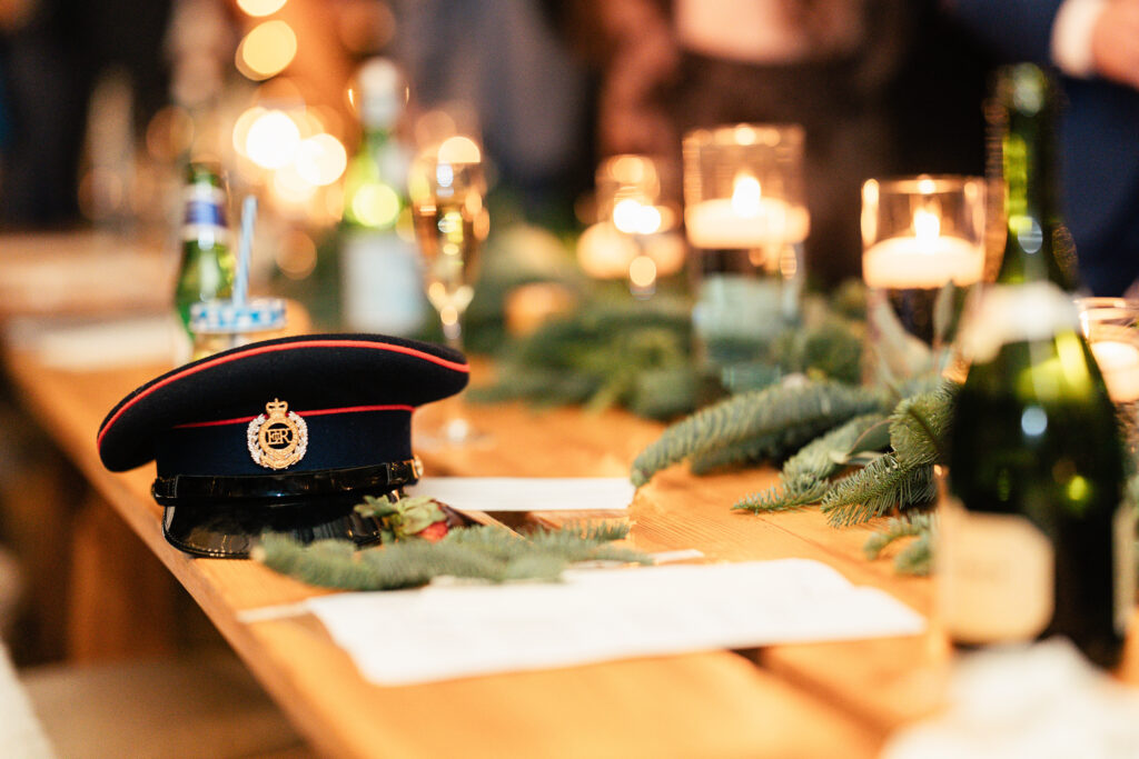 A wooden table with a uniform hat displaying a crest, surrounded by candles, greenery, and bottles. The setting suggests a festive or formal gathering, with soft, warm lighting in the background.