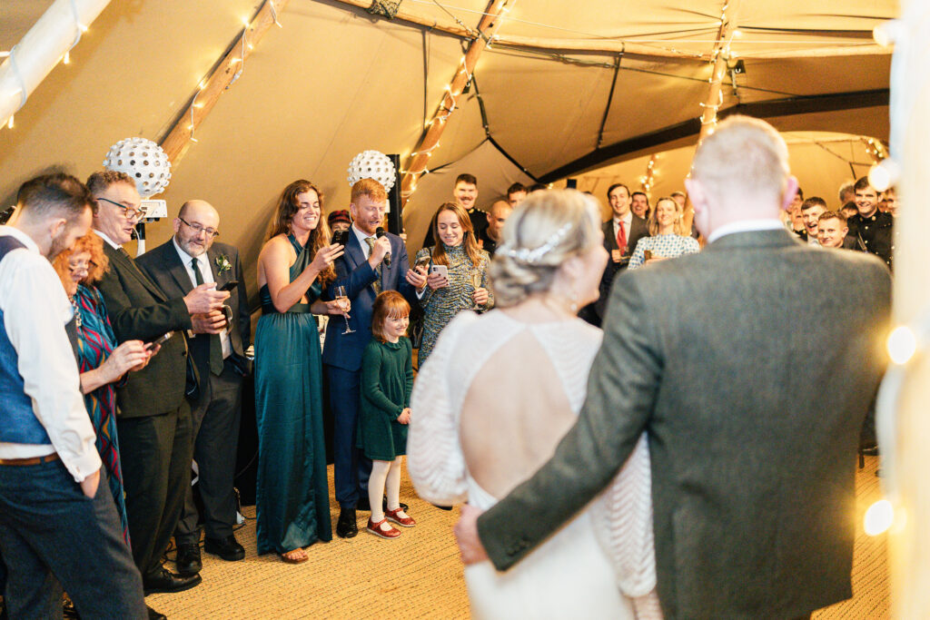 A couple stands embracing at the forefront, facing a group of people at a festive gathering. Guests, dressed in formal attire, watch and smile. The room is warmly lit with string lights, and a child in a green dress stands among the adults.