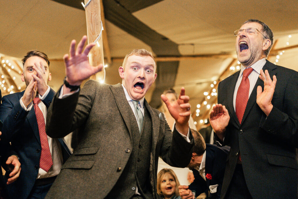 A group of people in suits with surprised and animated expressions. One man in a gray suit appears excited, while another in a dark suit and red tie is shouting. Twinkling lights are visible in the background, creating a festive atmosphere.