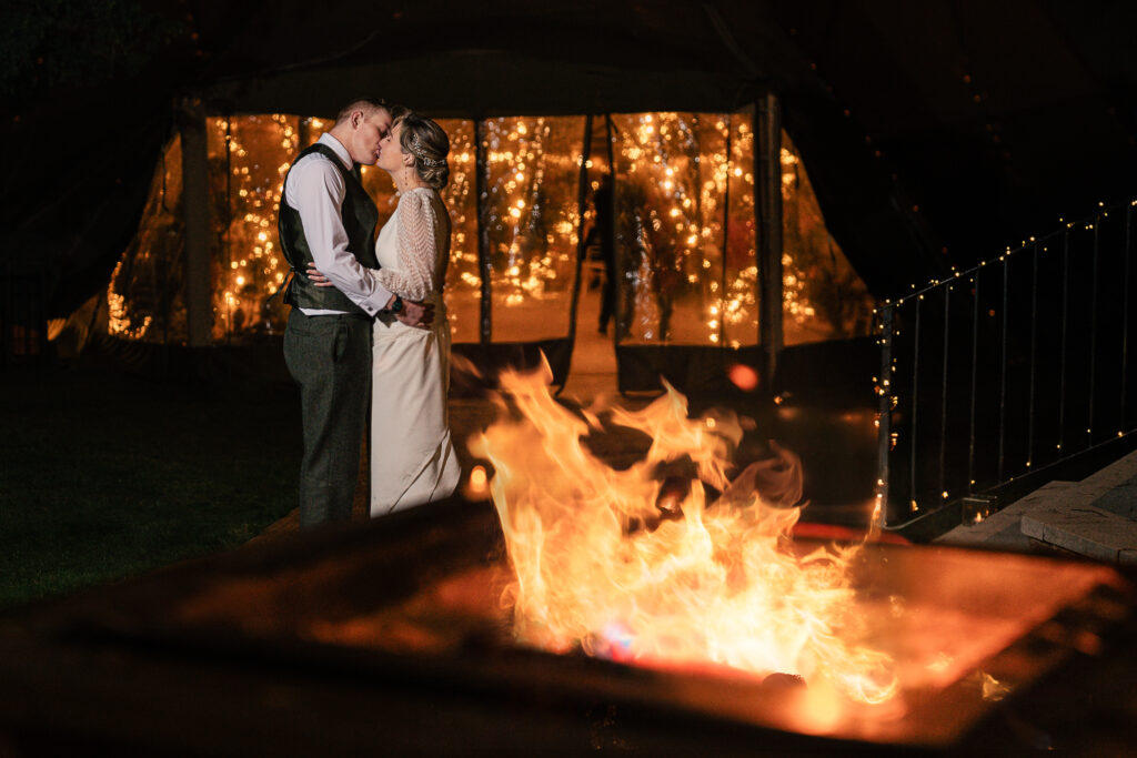 A couple embraces in front of a warmly lit tent, sharing a romantic moment. In the foreground, a fire pit glows with bright flames, adding to the cozy atmosphere of the evening scene.