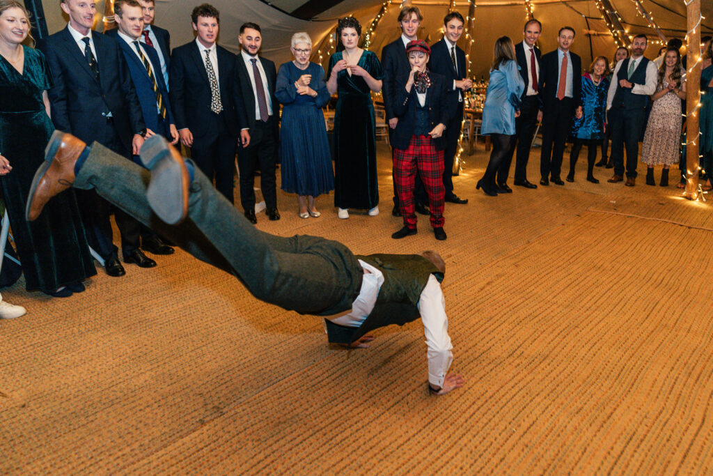 A person in formal attire performs a breakdance move on the floor while a crowd of people, dressed in suits and dresses, watches and smiles. The setting appears to be a festive indoor event with warm lighting and decorations.